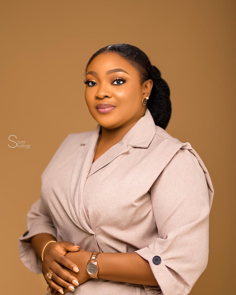 Professional headshot of a woman wearing a beige blazer against a tan background, with her hands clasped, smiling at the camera